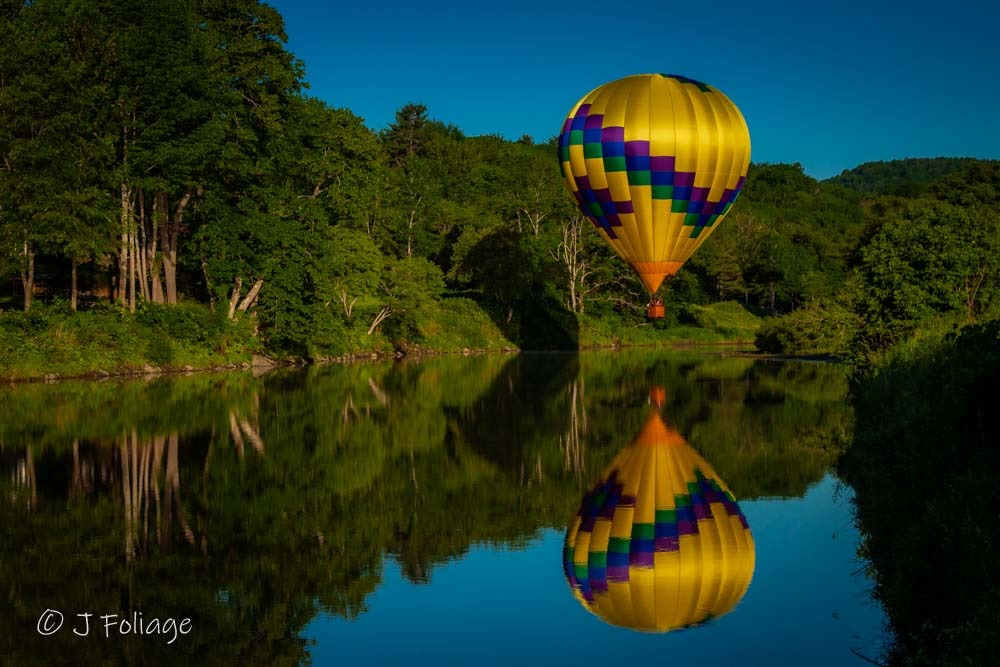 A yellow and purple patterned hot air balloon reflecting perfectly in the Ottauquechee River.