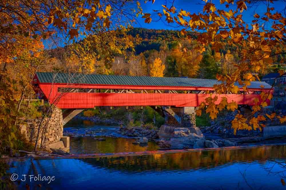 The red Taftsville Covered Bridge in Vermont surrounded by peak orange and yellow autumn foliage reflecting in the river.