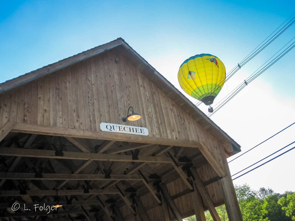 Colorful hot air balloons floating in a clear blue sky as seen from the pedestrian walkway of the historic Quechee Covered Bridge.