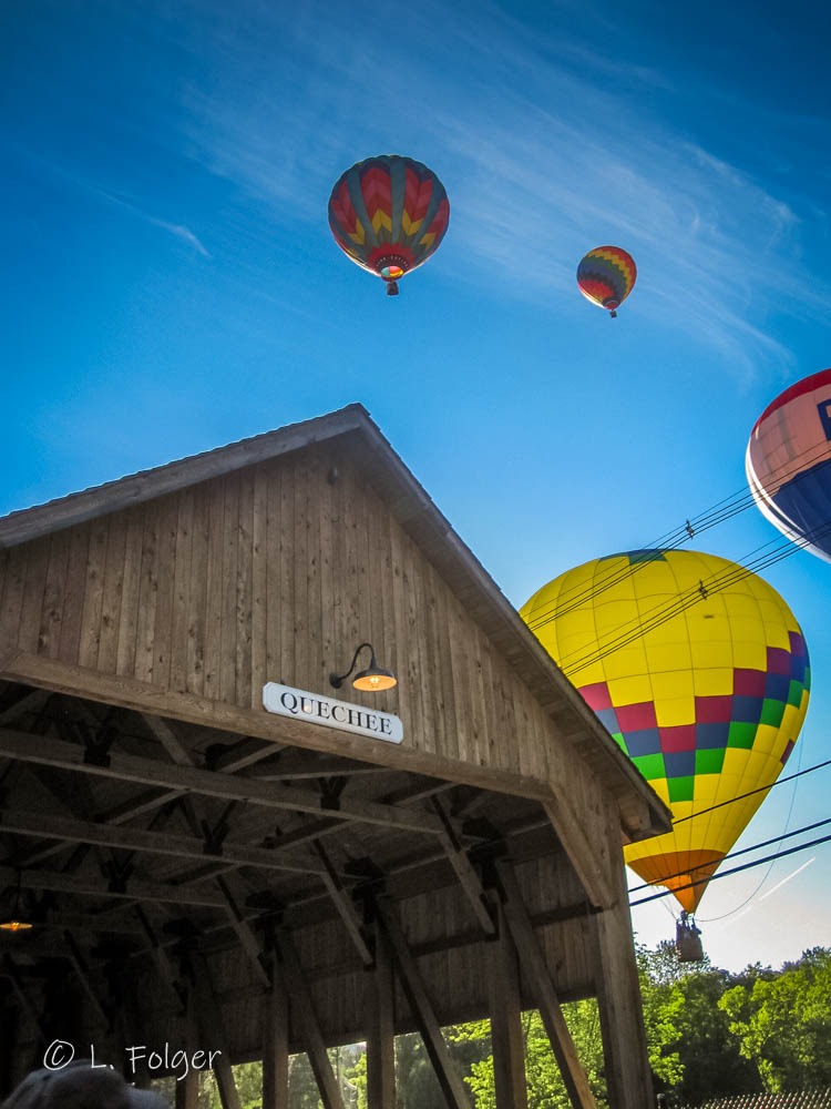 multiple Colorful hot air balloons floating in a clear blue sky as seen from the pedestrian walkway of the historic Quechee Covered Bridge.